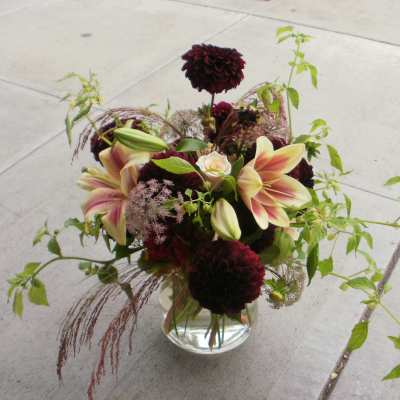 Low glass vase with burgundy dahlias, pink lilies, and trailing green vines.