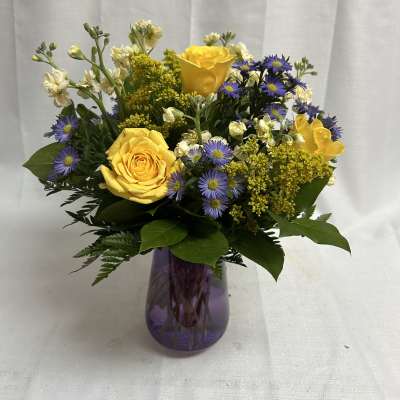 Yellow roses and purple daisies in a purple glass vase