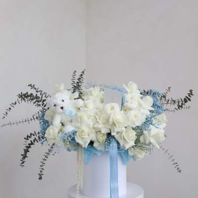 White rose bouquet with a teddy bear and blue ribbon in a white box