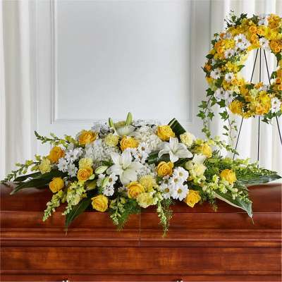 Yellow and white funeral casket spray with roses, lilies, and daisies, with matching wreath in background