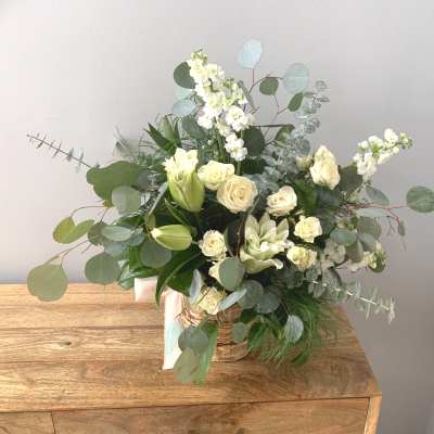 White and cream flower arrangement with roses and lilies in a woven basket with eucalyptus accents.