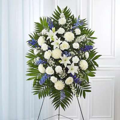 Tall standing spray of white lilies, roses, and other blooms with blue accents on a metal easel.