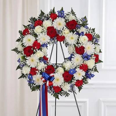 Patriotic standing wreath with red roses, white blooms, and blue flowers on an easel.