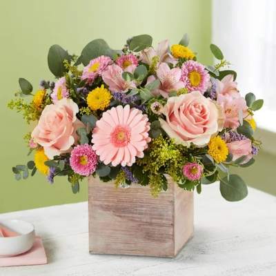 Low arrangement of pink roses, gerbera daisies, and yellow button blooms in a rustic wooden cube vase.