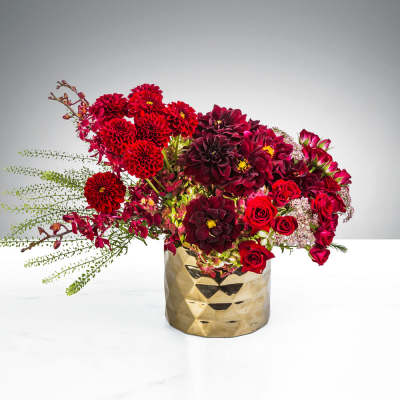 Asymmetrical arrangement of red and burgundy dahlias and roses in a gold textured vase