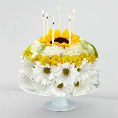 Round floral cake of white daisies, yellow blooms, and a sunflower on a stand with six lit candles on top.