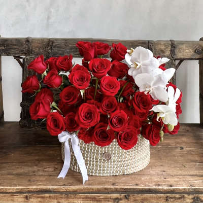 Red roses and white orchids arranged in a woven basket with a white ribbon