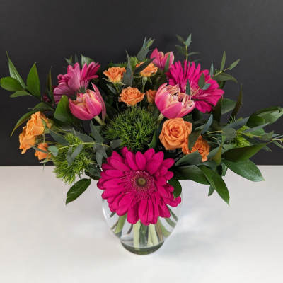 Bright arrangement of hot pink gerbera daisies, pink tulips, and orange roses in a clear glass vase