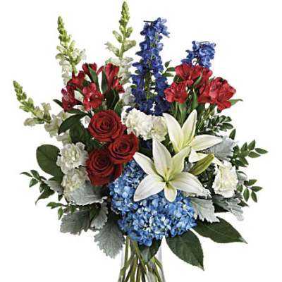 Tall red, white, and blue flower arrangement with roses, lilies, and delphinium in a clear glass vase