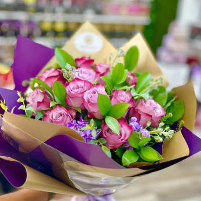 Handheld bouquet of pink roses with purple accents wrapped in tan and purple paper with a ribbon