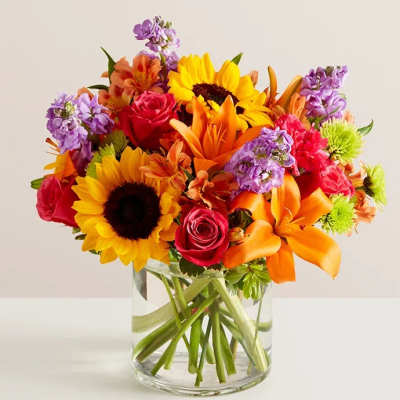 Mixed bouquet of sunflowers, roses, lilies, and purple blooms in a glass vase