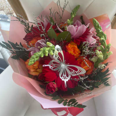 Bright bouquet of red, pink, and orange flowers wrapped in pink paper with a silver butterfly decoration.