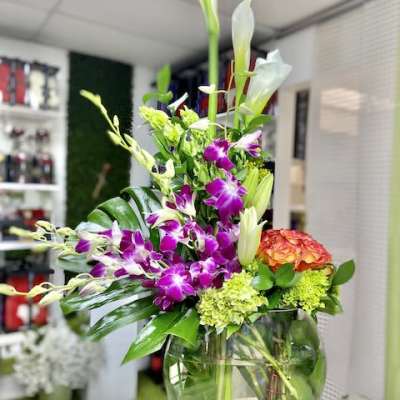 Tall mixed arrangement with white calla lilies, purple orchids, and orange blooms in a round glass vase