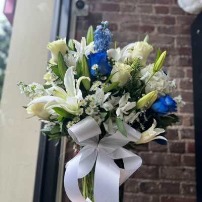 Tall arrangement of white lilies, roses, and blue accent flowers in a glass vase with a white bow