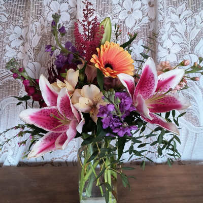 Mixed bouquet with pink lilies, orange gerbera daisy, and purple flowers in a glass vase