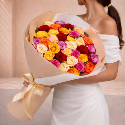 Large bouquet of multicolored roses wrapped in beige paper and held by a woman in a white dress.