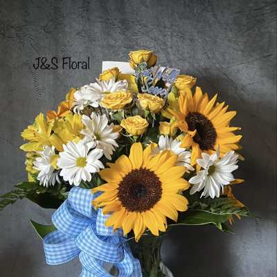 Bouquet of sunflowers, white daisies, and yellow roses in a glass vase with a blue ribbon
