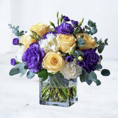 Yellow roses and purple carnations in a clear square vase