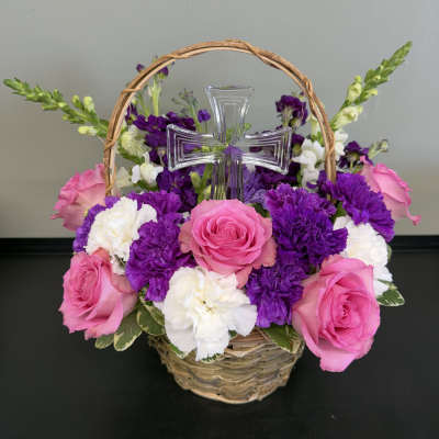 Basket arrangement of pink roses, white carnations, and purple flowers with a clear cross