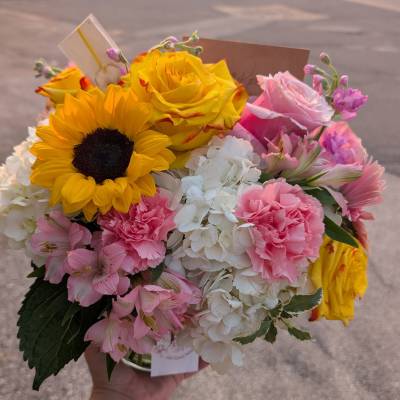 Round mixed bouquet with sunflower, yellow roses, pink carnations, and white hydrangeas in a clear vase
