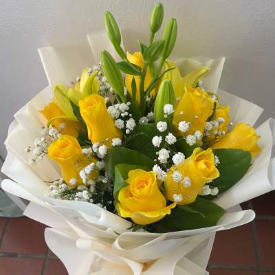 Hand-tied bouquet of yellow roses, lily buds, and white filler flowers wrapped in white paper.