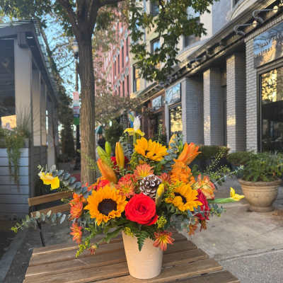 Autumn arrangement with sunflowers, orange lilies, and red roses in a cream vase on a wooden table.