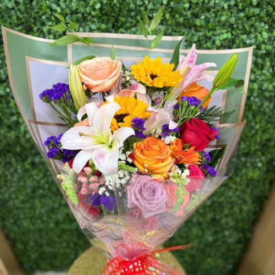 Colorful wrapped bouquet with lilies, roses, sunflowers, and mixed blooms tied with a red ribbon