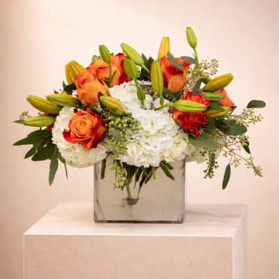 Low glass vase arrangement with orange roses, white hydrangeas, and green lily buds.