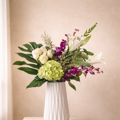 Tall white vase with white roses, lilies, green hydrangea and purple orchids with large tropical leaves.