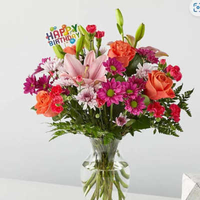 Birthday bouquet with pink lilies, orange roses, and purple daisies in a clear glass vase