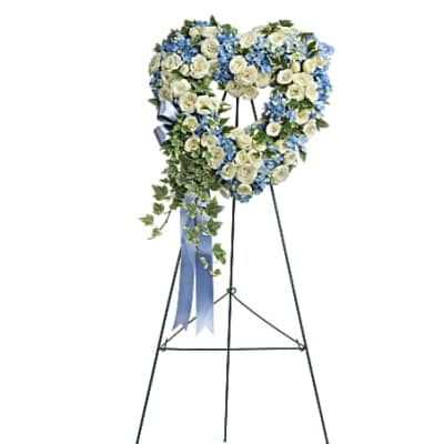 Heart-shaped standing wreath of white roses and blue flowers with ivy cascade and blue ribbon on a black easel