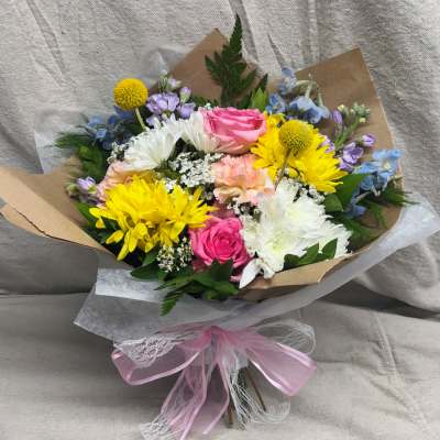 Hand-tied bouquet of pink roses, yellow daisies, and white mums wrapped in kraft paper with a pink bow.
