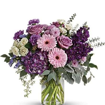 Medium arrangement of pink gerbera daisies, lavender roses, and purple blooms in a clear glass vase