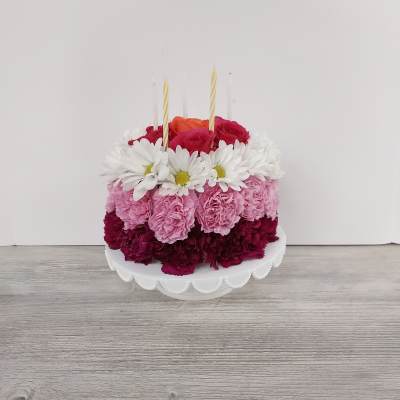 Round floral cake of pink carnations, white daisies, and roses with candles on a white cake stand