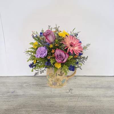 Colorful mixed flower arrangement with pink gerbera, roses, and purple blooms in a floral ceramic mug