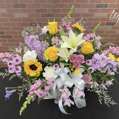 Mixed floral arrangement with yellow roses, lilies, sunflowers, and pink blooms in a white basket