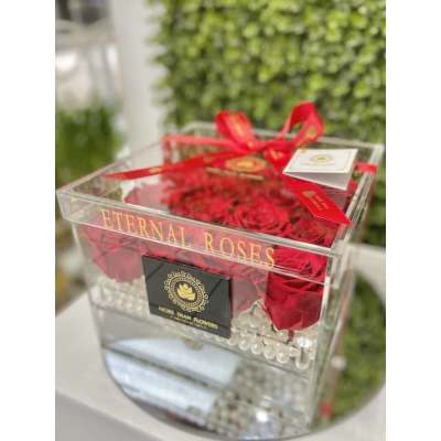 Clear acrylic box filled with red roses and pearl accents, tied with a red ribbon and card.