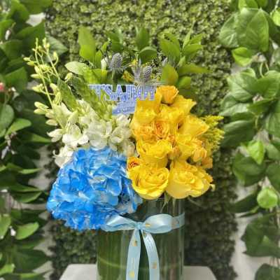 Tall glass vase with yellow roses, blue hydrangea, white orchids and an 'It’s a Boy' pick.