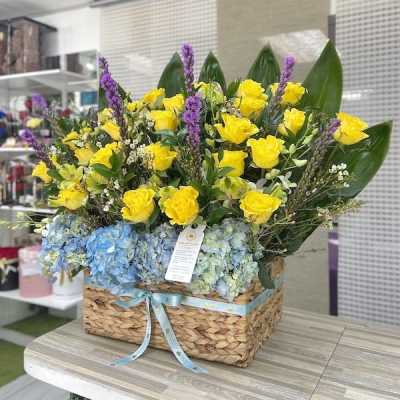 Large basket of yellow roses, blue hydrangeas, purple flowers and bold green leaves with a blue ribbon.