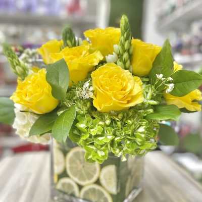 Square glass vase filled with lemon slices holding yellow roses and green hydrangeas with white accents.