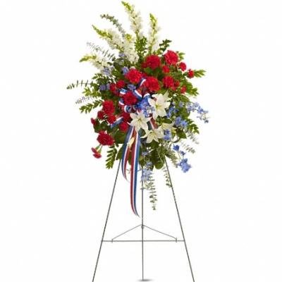 Patriotic standing spray of red carnations, white lilies, and blue flowers on a metal easel.