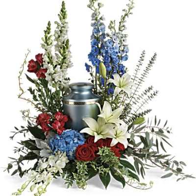 Cremation urn surrounded by red roses, white lilies, blue hydrangea, and other mixed flowers.