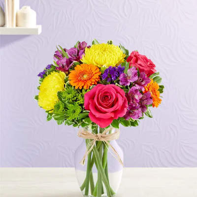 Bright mixed bouquet of pink roses, yellow mums, orange gerberas and purple blooms in a clear glass vase.