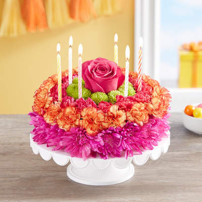 Round floral cake arrangement with pink, orange, and green flowers topped with candles on a white stand