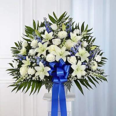 Large white and blue floral arrangement with lilies, roses, and ribbon on a white pedestal.