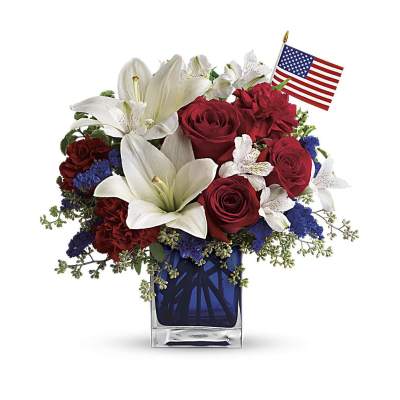Red, white, and blue flower arrangement with lilies and roses in a blue vase with a small U.S. flag.