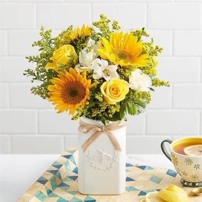 Yellow sunflowers, roses, and white blooms in a tall white ceramic vase with a bow