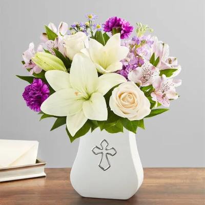 White lilies, roses, and purple flowers in a white vase with a silver cross design.