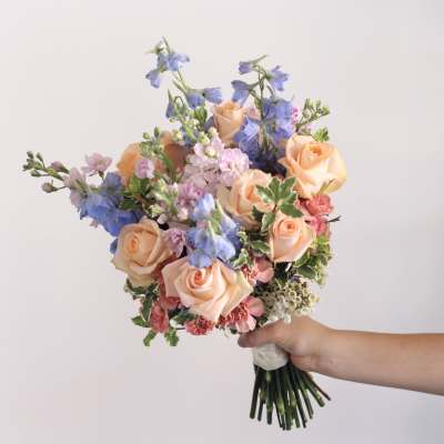 Handheld bouquet of peach roses, blue delphinium, and pink flowers