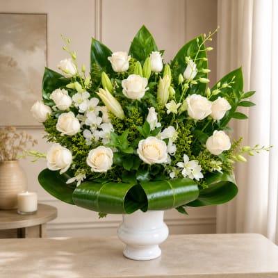 White roses and orchids arranged in a white vase with broad green leaves.